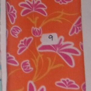 May include: A rectangular piece of fabric featuring a vibrant orange background with a repeating pattern of pink flowers and thin yellow stems. A small white rectangular label with the handwritten number 9 is attached to the center of the fabric.