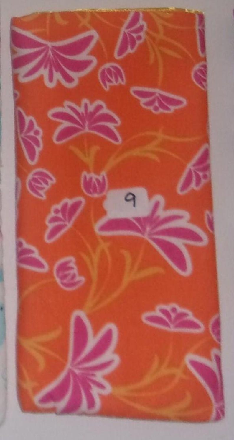 May include: A rectangular piece of fabric featuring a vibrant orange background with a repeating pattern of pink flowers and thin yellow stems. A small white rectangular label with the handwritten number 9 is attached to the center of the fabric.