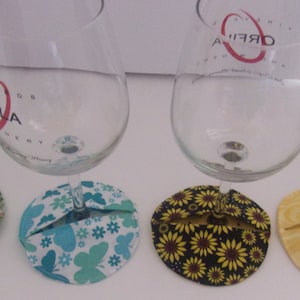 May include: Four wine glasses with decorative fabric coasters. The glasses have various designs, including a ladybug and winery logos. The coasters feature floral and butterfly patterns in green, blue, yellow, and black.