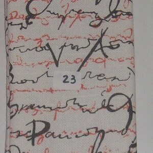May include: A rectangular fabric case featuring an off-white background with a pattern of black cursive handwriting layered over horizontal red dashed lines. A small white rectangular label with the number 23 is centered on the front surface.