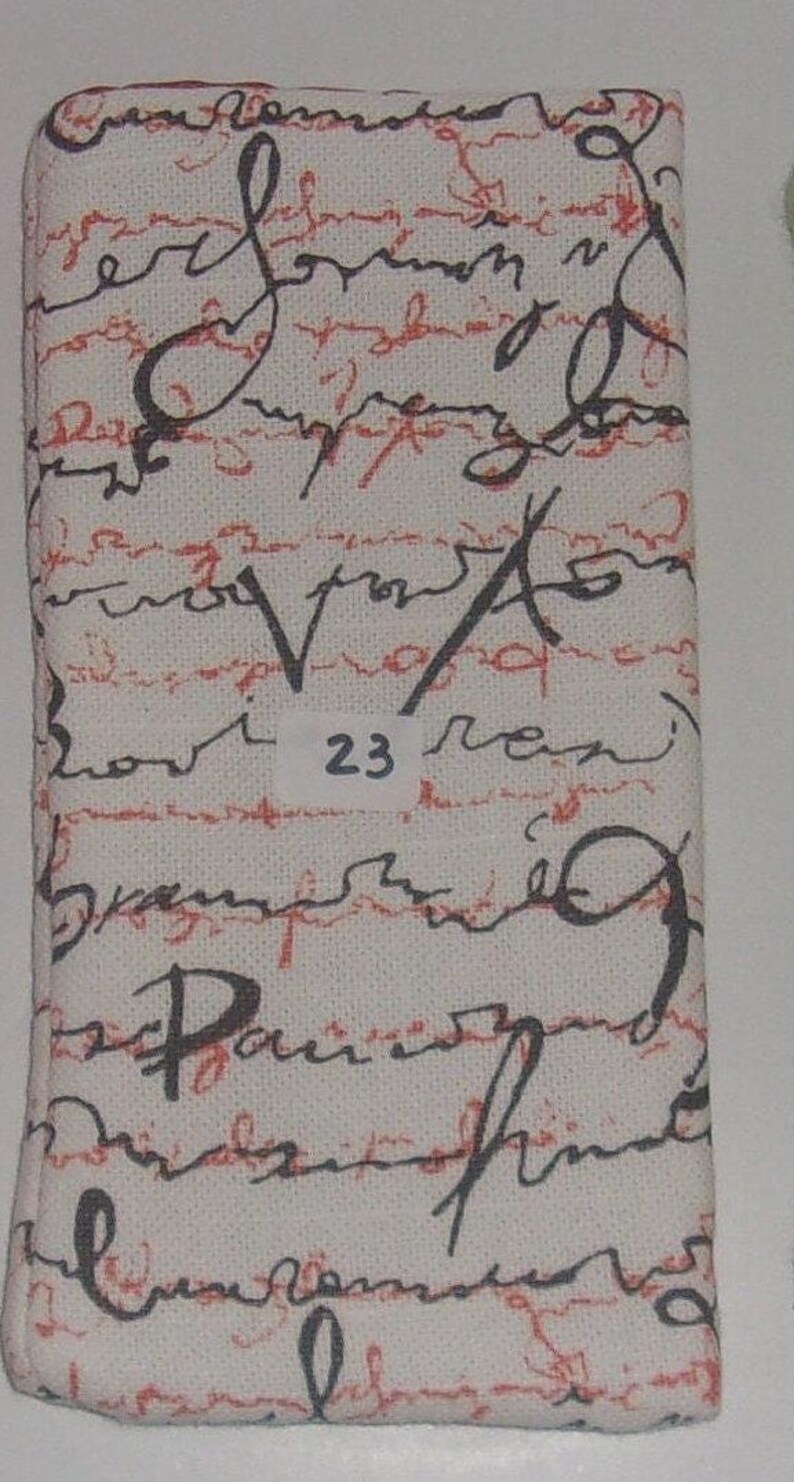 May include: A rectangular fabric case featuring an off-white background with a pattern of black cursive handwriting layered over horizontal red dashed lines. A small white rectangular label with the number 23 is centered on the front surface.