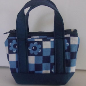 May include: A small, navy blue tote bag with a floral and checkered pattern in shades of blue and white. The bag has a zipper closure and navy blue handles. The base of the bag is a solid navy blue.