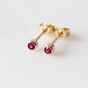 May include: A pair of gold stud earrings featuring small, round, ruby-red gemstones. The earrings are displayed on a white surface, highlighting their delicate design and vibrant colour. These earrings are a classic jewellery item.