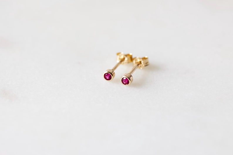 May include: A pair of gold stud earrings with small, round, ruby-red gemstones. The earrings are set on a white surface, showcasing their delicate design and vibrant color. The earrings are a classic jewelry item.