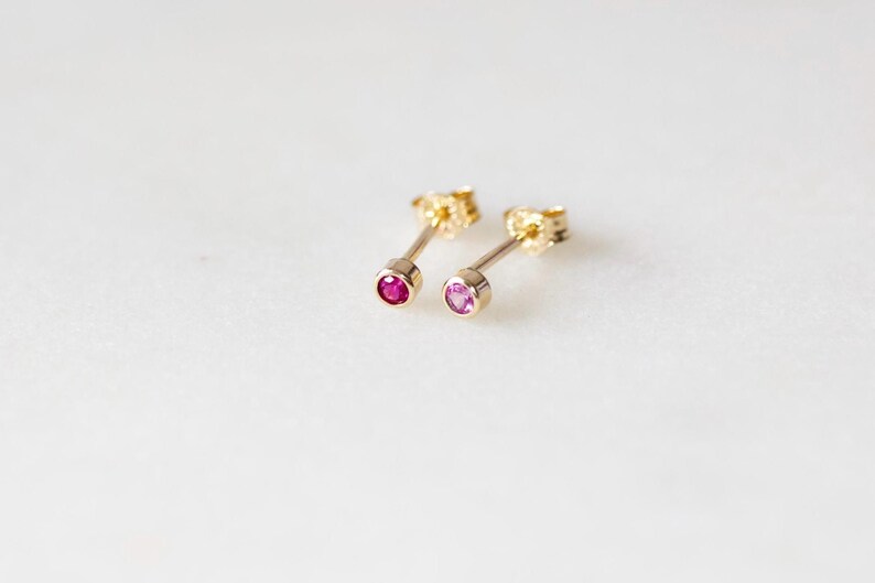 May include: Two gold stud earrings, each with a small, round gemstone. One earring features a deep red gemstone, while the other has a lighter pink stone. The earrings are set against a white background.