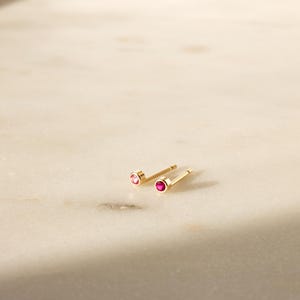 May include: A pair of gold stud earrings with small, round, red gemstones. The earrings are displayed on a light-colored, marble-like surface. The studs are simple and elegant, suitable for everyday wear.