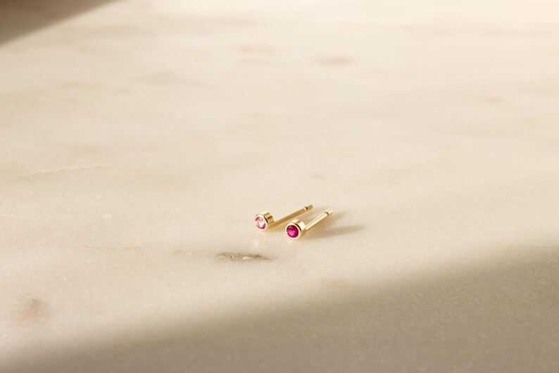 May include: A pair of gold stud earrings with small, round, red gemstones. The earrings are displayed on a light-colored, marble-like surface. The studs are simple and elegant, suitable for everyday wear.