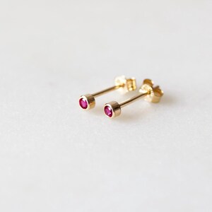 May include: A pair of gold stud earrings with small, round, ruby-colored gemstones. The earrings are set on a white surface, showcasing their delicate design and vibrant color. The earrings are a classic jewelry item.