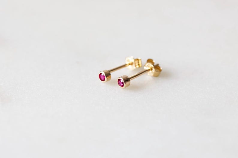 May include: A pair of gold stud earrings with small, round, ruby-colored gemstones. The earrings are set on a white surface, showcasing their delicate design and vibrant color. The earrings are a classic jewelry item.