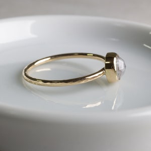 May include: A gold ring with a hammered band and a clear, faceted gemstone. The ring is on a white ceramic dish.