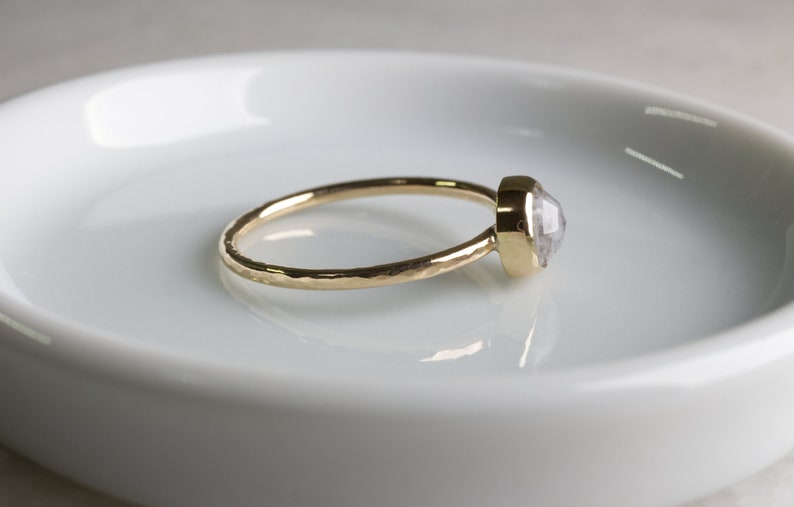 May include: A gold ring with a hammered band and a clear, faceted gemstone. The ring is on a white ceramic dish.