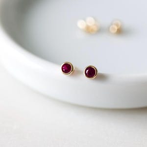 May include: A pair of gold stud earrings with round, deep red gemstones. The earrings are set in a gold bezel and sit on a white, curved surface. Additional gold earrings are blurred in the background.