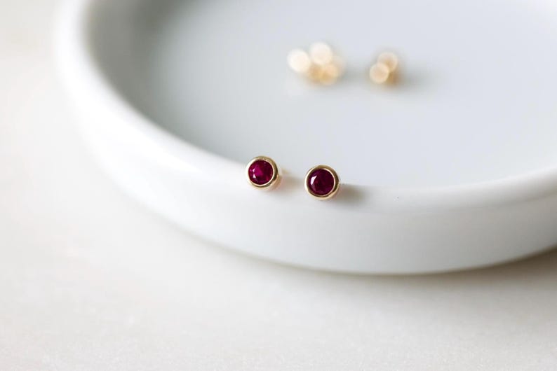 May include: A pair of gold stud earrings with round, deep red gemstones. The earrings are set in a gold bezel and sit on a white, curved surface. Additional gold earrings are blurred in the background.