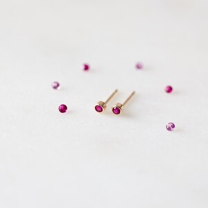 May include: A pair of gold stud earrings with small, round, ruby-colored gemstones. Several loose, similar gemstones are scattered around the earrings on a white surface. The earrings have a simple, minimalist design.