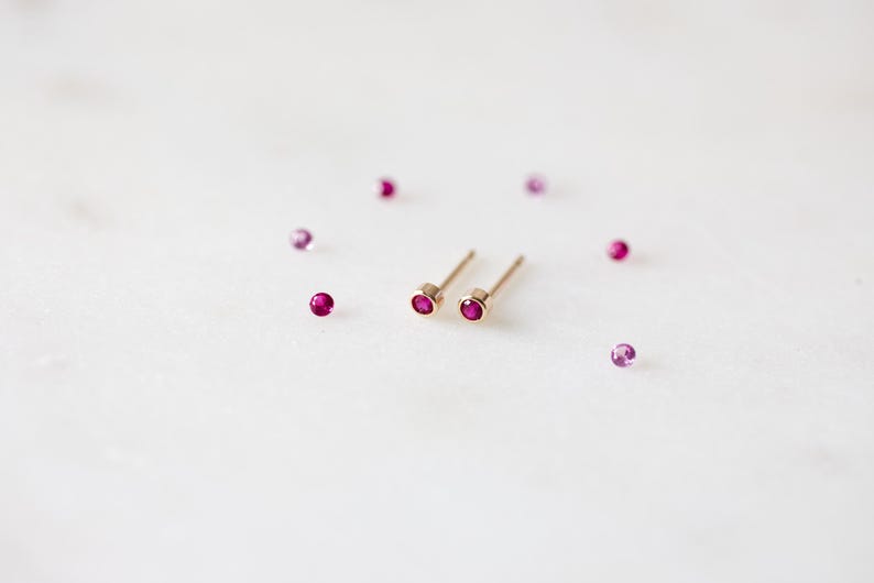 May include: A pair of gold stud earrings with small, round, ruby-colored gemstones. Several loose, similar gemstones are scattered around the earrings on a white surface. The earrings have a simple, minimalist design.