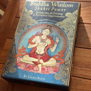 May include: A blue box with gold detailing, titled "Buddha Wisdom Shakti Power." The box displays an illustration of a seated figure in red and orange attire, within a blue circle. The text "By Laura Santi" is also present.