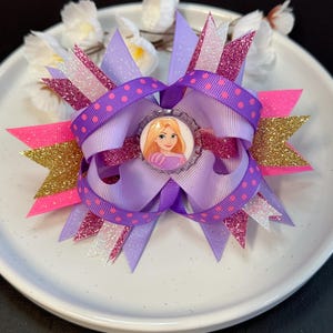 May include: Purple, pink, and gold glitter hair bow with a purple polka dot ribbon and a round, clear button featuring a cartoon image of Rapunzel from the Disney movie Tangled.