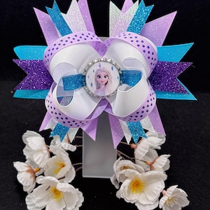 May include: A multi-layered hair bow featuring purple, white, and blue ribbons with glitter and polka dot patterns. A circular bottle cap center displays a graphic of a blonde character with blue eyes. White artificial flowers are attached below.
