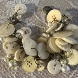 May include: A cluster of off-white buttons and faux pearls are connected by a silver-colored chain. The buttons vary in size and shape, with some having four holes and others two. The pearls are small and round, adding a touch of elegance to the design.