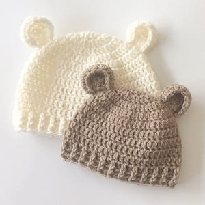 Può includere: Due cappelli all'uncinetto con orecchie da orso. Un cappello è bianco e l'altro è marrone.