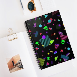 May include: A black spiral notebook with a colorful geometric pattern featuring aliens, planets, and rockets. The pattern is in neon colors like pink, green, blue, and yellow.