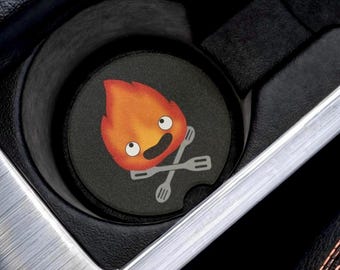 May include: A black car cup holder with a round, black coaster featuring a cartoon fire spirit with a smiling face and crossed forks. The fire spirit is orange and yellow.