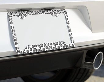 May include: A white and gray leopard print license plate frame. The frame is made of metal and has a shiny finish.