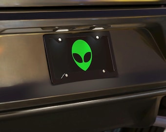 May include: A black rectangular license plate frame featuring a bright green alien head graphic centered on the plate. The frame is mounted to the rear bumper of a vehicle with four silver screws visible at the corners of the frame.