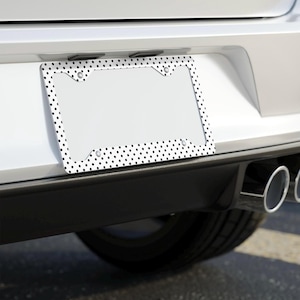 May include: A white car with a black and white polka dot license plate frame. The frame is made of metal and has a raised edge around the perimeter. The frame is attached to the car's bumper with screws.