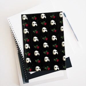 May include: A black notebook with a repeating pattern of white Phantom of the Opera masks and red roses.