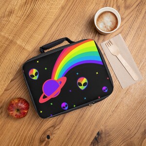 May include: Black lunch bag with a colorful rainbow, planet, and alien design. The bag has a black handle and zipper. A red apple, a cup of coffee, and a wooden fork are on a wooden surface.