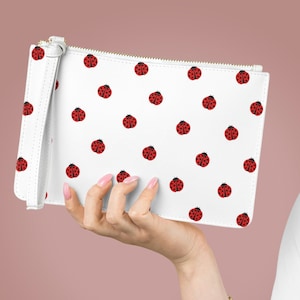 May include: A white clutch purse with a zipper closure and a wrist strap. The purse is covered in a repeating pattern of red ladybugs.