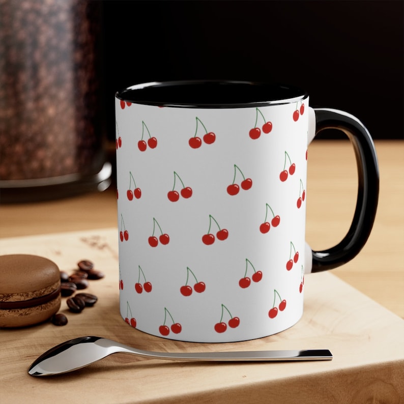 May include: White ceramic mug with a black rim and handle. The mug features a repeating pattern of red cherries on a white background.