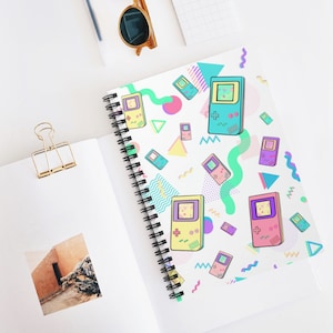 May include: A white spiral notebook with a colorful pattern of retro video game consoles and geometric shapes. The pattern includes pink, blue, green, yellow, and purple colors.