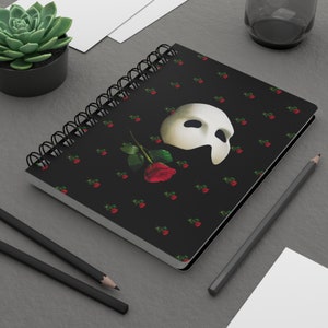 May include: Spiral-bound notebook with a black cover featuring a white mask, a red rose, and small red rose patterns. The notebook is on a gray surface with pencils and a succulent plant.
