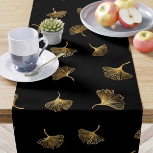 May include: Black table runner with a gold ginkgo leaf pattern. The runner is on a wooden table with a white plate, a cup, and a small potted plant.