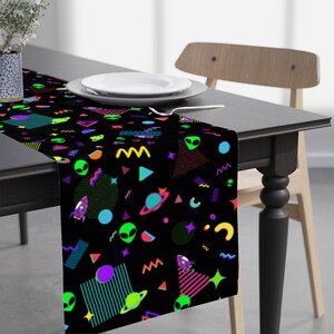 May include: A black table runner with a colorful geometric and alien pattern. The pattern includes neon green, pink, blue, and yellow colors. The runner is on a black table with a white plate and a silver fork.