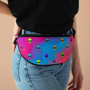 May include: A colorful fanny pack with a vibrant gradient background of pink, blue, and purple. The bag is adorned with multiple alien head graphics in various colors. The fanny pack has a black zipper and a black strap.
