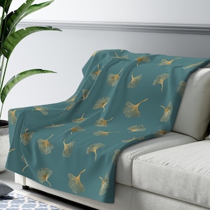 May include: A teal blue throw blanket with a pattern of gold ginkgo leaves. The blanket is draped over a light gray couch.