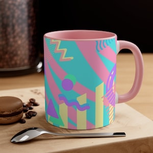 May include: A pink ceramic mug with a geometric pattern in shades of blue, pink, yellow, and purple. The pattern includes stripes, circles, and wavy lines.