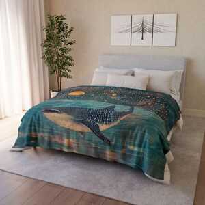 May include: A teal blue and gold whale blanket with a starry night sky design. The whale is swimming through the water with a full moon in the background.