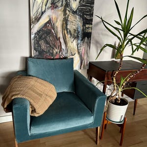 May include: A teal velvet armchair with wooden legs and a brown knitted throw blanket. The chair is in front of a wall with a large abstract painting and a potted plant.