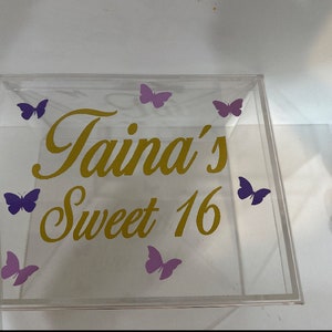 May include: A clear square acrylic box featuring gold cursive text that reads Taina's Sweet 16. The lid is decorated with six purple butterfly decals arranged around the lettering. The box has a transparent construction with a flat top surface.