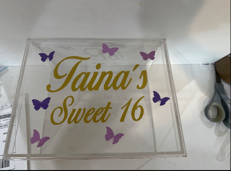 May include: A clear square acrylic box featuring gold cursive text that reads Taina's Sweet 16. The lid is decorated with six purple butterfly decals arranged around the lettering. The box has a transparent construction with a flat top surface.