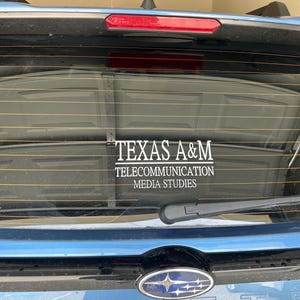 May include: A blue car's rear window displays a white decal with the text "TEXAS A&M" above "TELECOMMUNICATION MEDIA STUDIES." The window has a wiper and a red brake light. The car's emblem is visible.