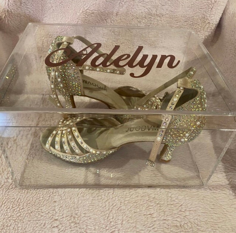 May include: A pair of champagne-colored heels with an open-toe design, covered in clear crystals and small white pearls, enclosed in a clear acrylic box with the name Adelyn printed in dark brown script on the front surface of the container.