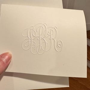 May include: A white card with a raised, embossed monogram of the letters "SBR" in a cursive script.