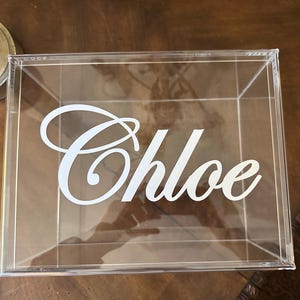 May include: A clear, square acrylic box featuring the name Chloe in white, cursive lettering applied to the front panel. The container is transparent, allowing for a view through the structure, and is constructed with clean, sharp edges.