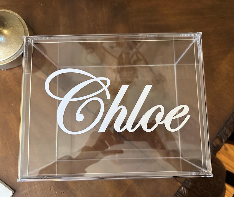 May include: A clear, square acrylic box featuring the name Chloe in white, cursive lettering applied to the front panel. The container is transparent, allowing for a view through the structure, and is constructed with clean, sharp edges.
