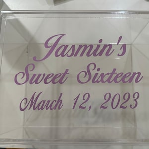 May include: A clear, square acrylic box featuring purple cursive text printed on the front surface. The text reads: Jasmin's Sweet Sixteen, March 12, 2023. The box is transparent, allowing a view through the material to the interior and the opposite side.